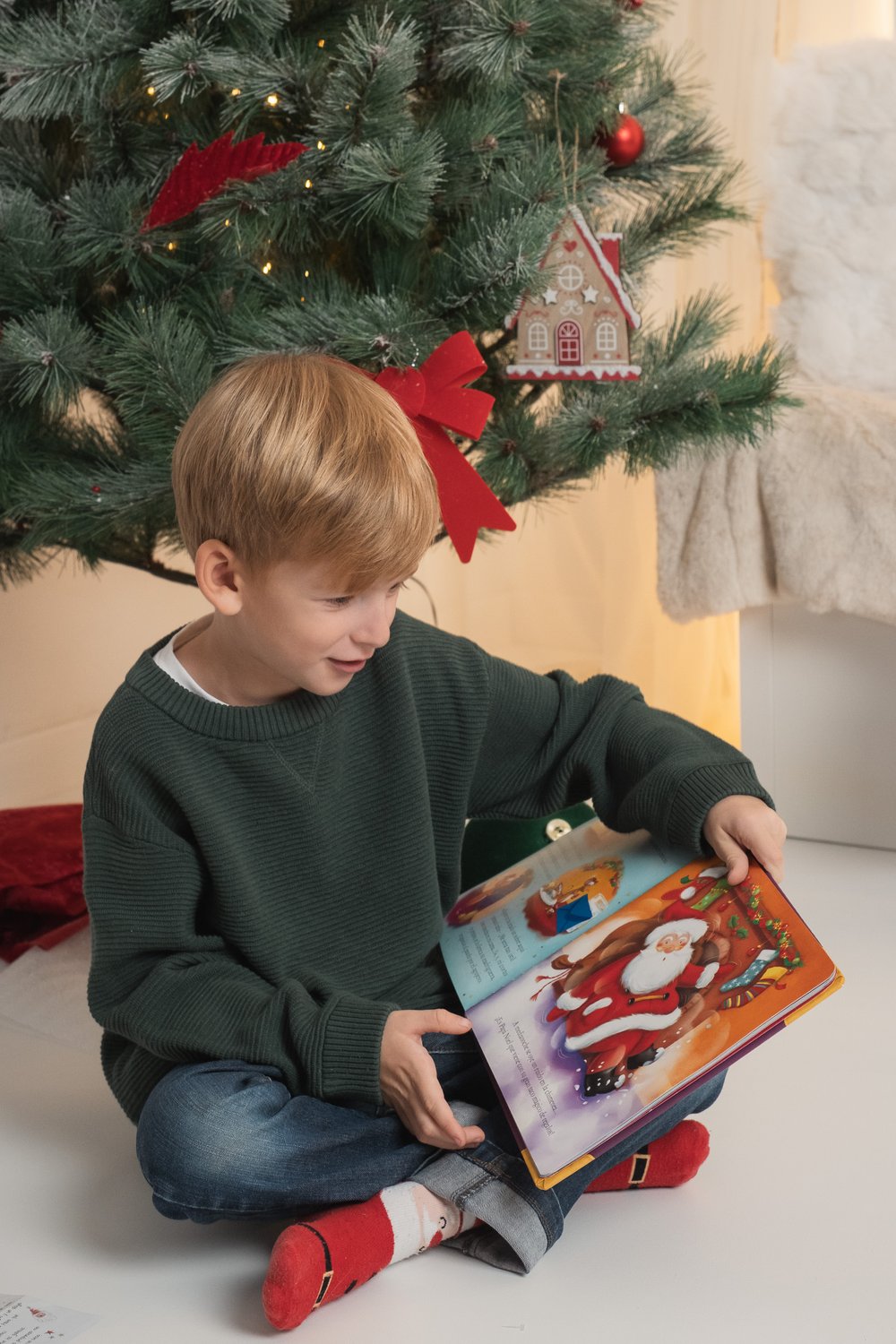 Niño sonriendo con un cuento bajo el árbol en una sesión de fotos navideña en familia