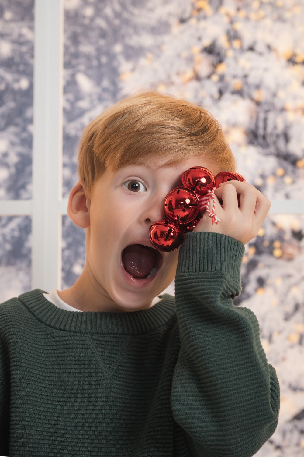 Sesión de fotos de Navidad en Alicante: niño con cascabeles rojos junto a una ventana nevada